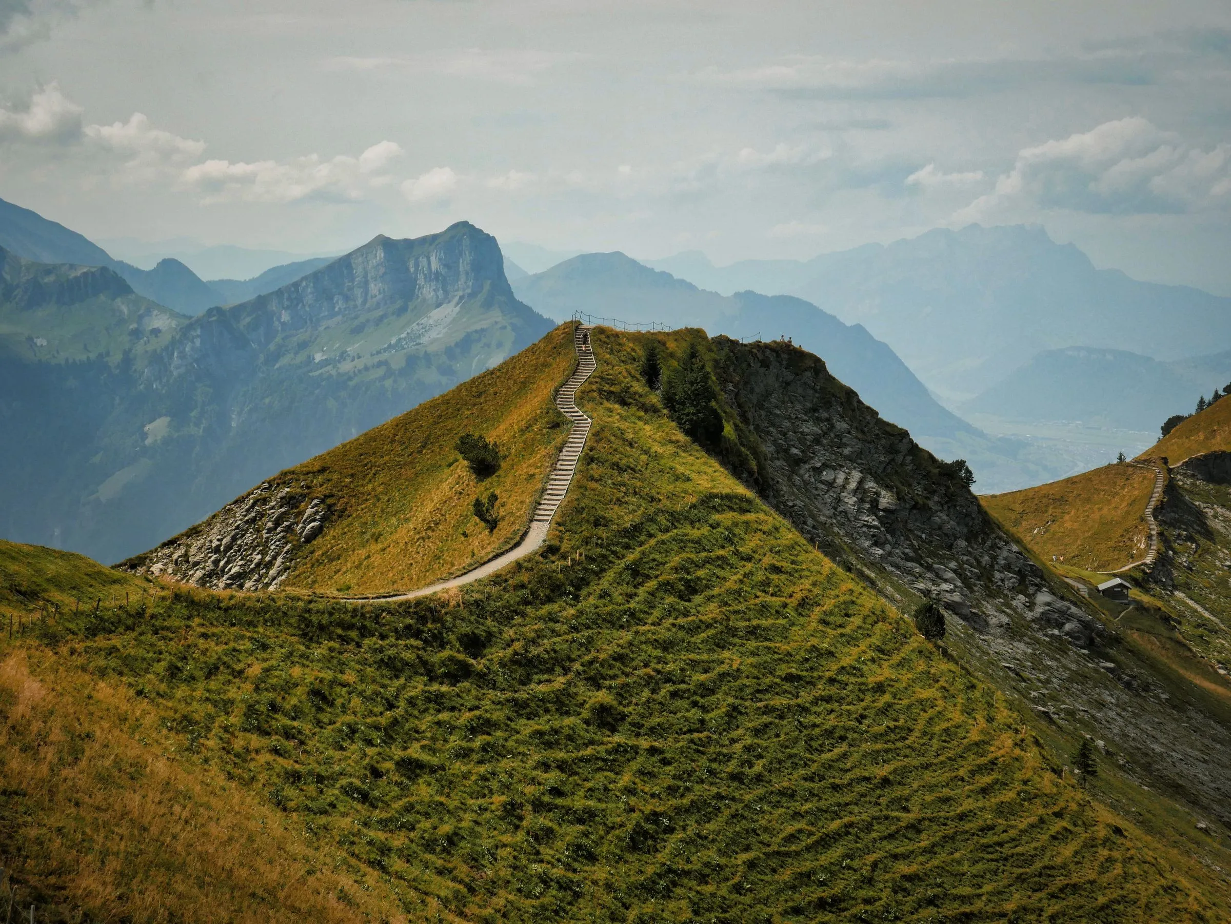 A grassy ridge with a worn path curving along its crest, steep slopes falling away on both sides under a hazy sky