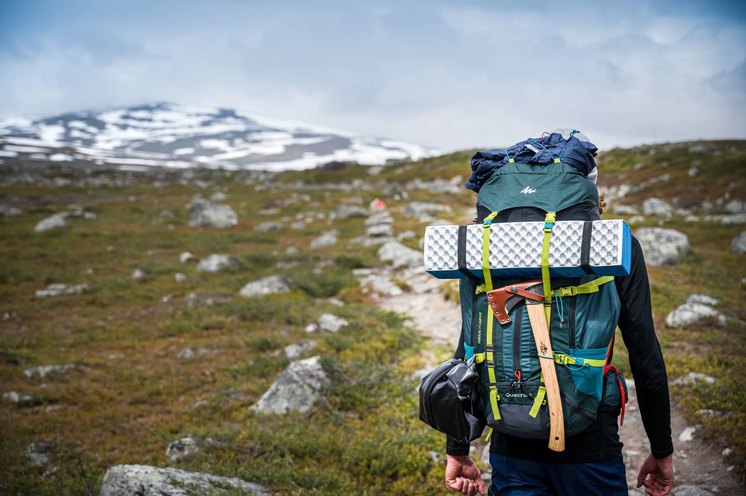 A walker seen from behind carrying a full backpack with a foam mat strapped on, heading up a stony path towards snow-streaked hills