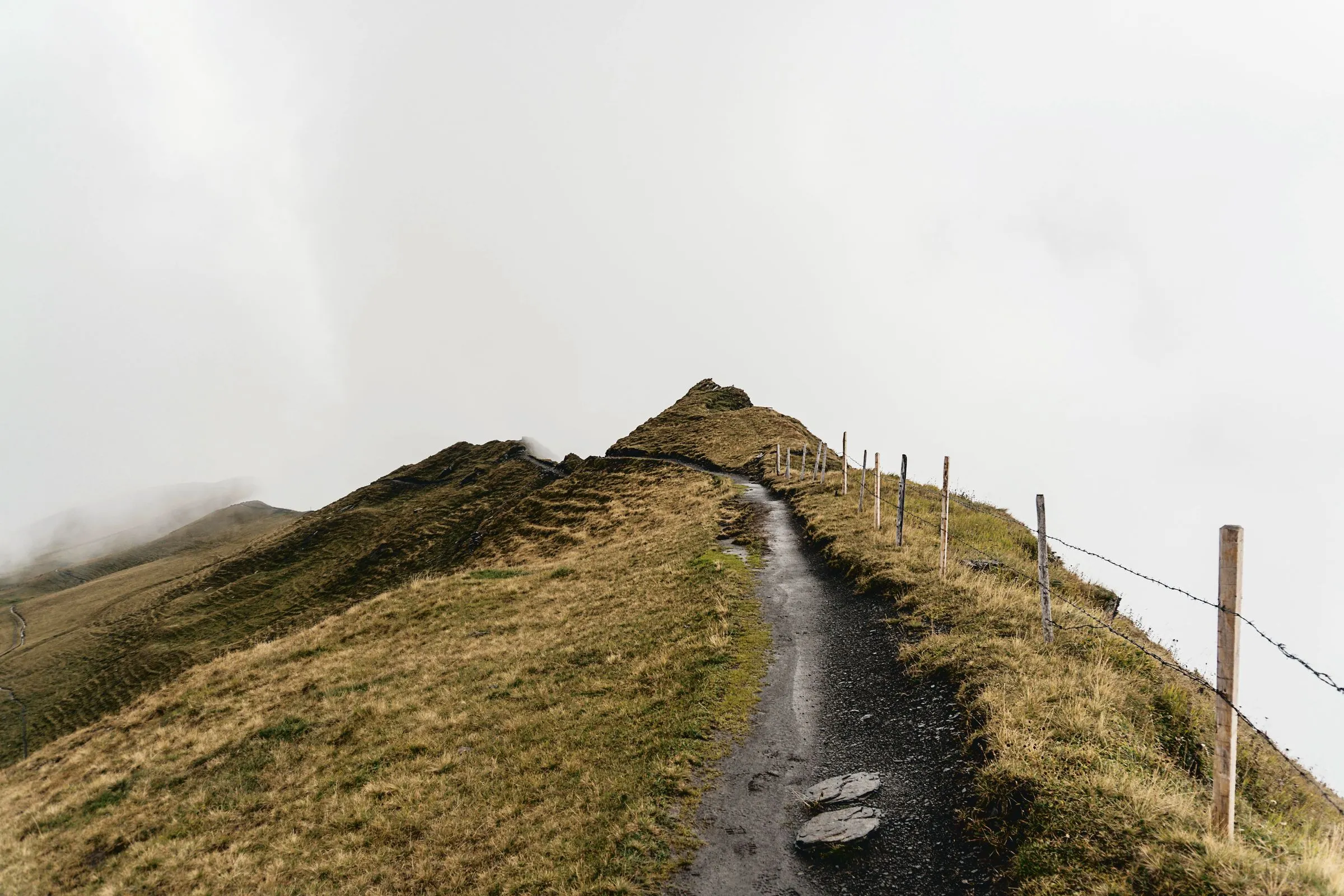A wet path climbing a grassy hill beside a wire fence and wooden posts, the top lost in thick grey cloud
