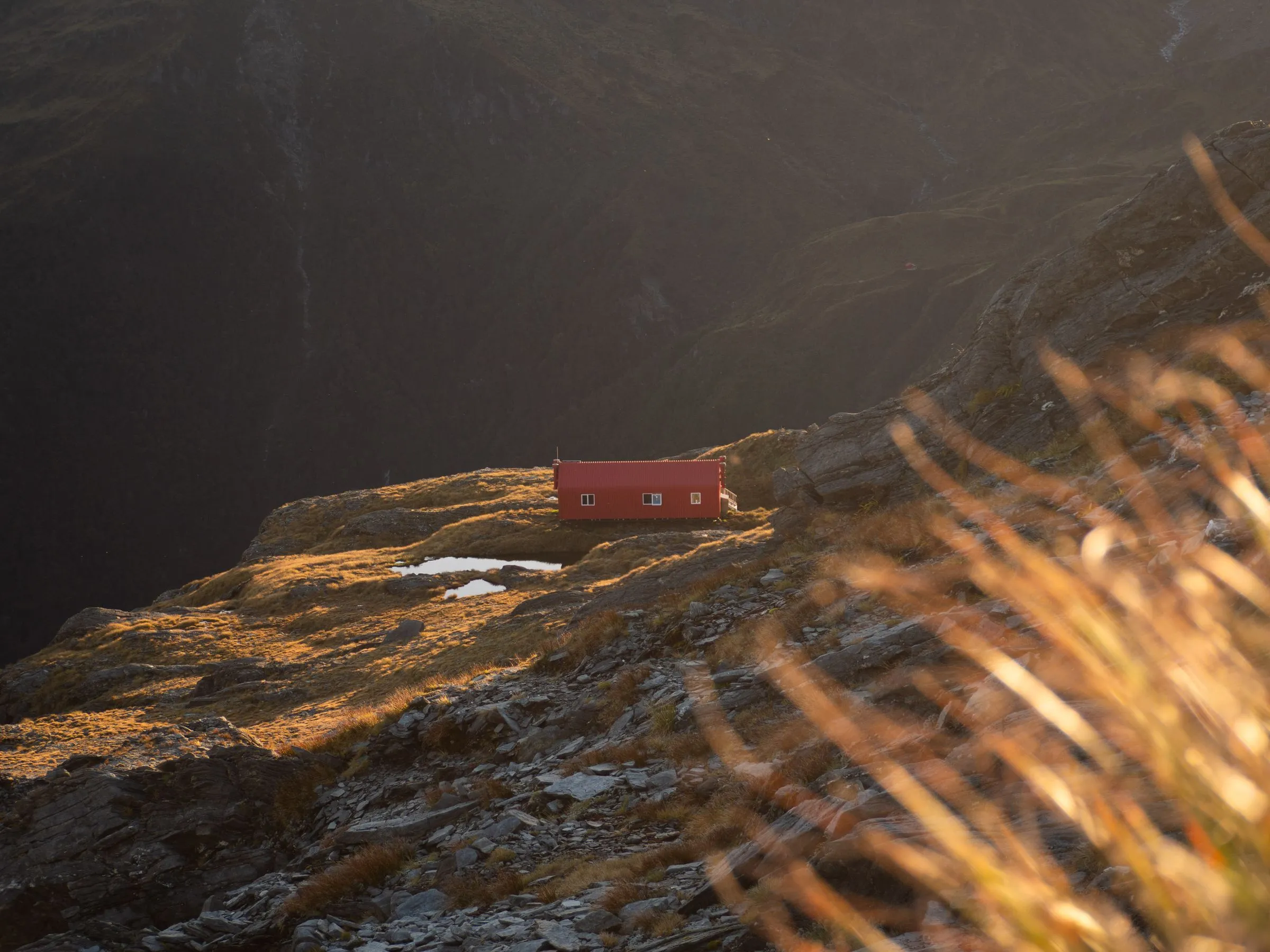 A small red hut on a high grassy shoulder, lit orange by the evening sun, with mountains in shadow behind
